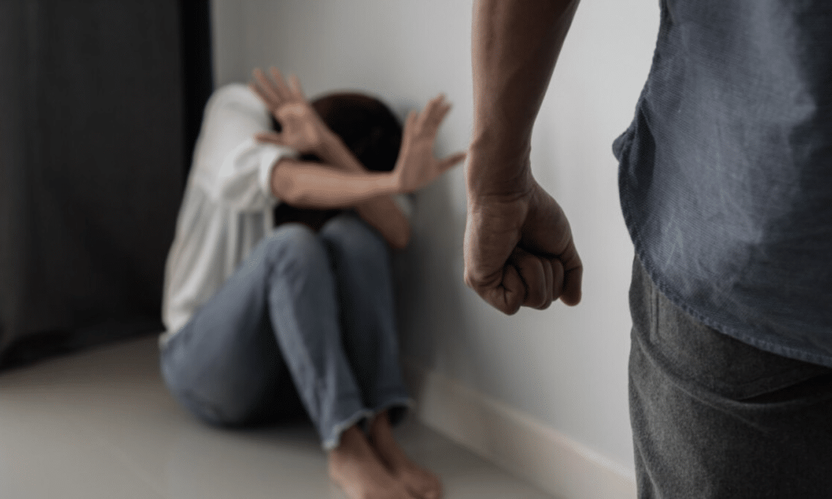 Чоловік із стиснутим кулаком стоїть навпроти наляканої людини, яка сидить у кутку та закривається руками.