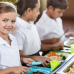 Усі школярі з вересня отримуватимуть безкоштовне харчування Школярі у шкільній їдальні отримують гарячий обід — діти стоять біля лінії роздачі з тарілками їжі.