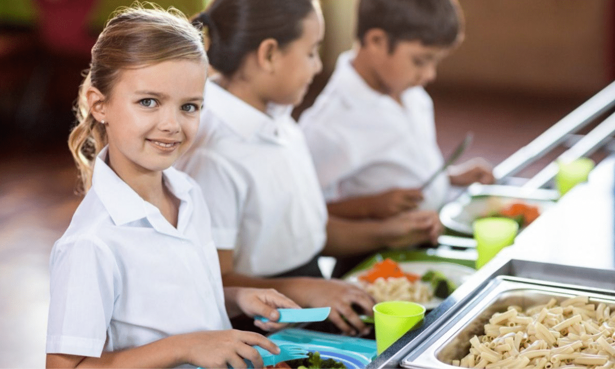 Школярі у шкільній їдальні отримують гарячий обід — діти стоять біля лінії роздачі з тарілками їжі.