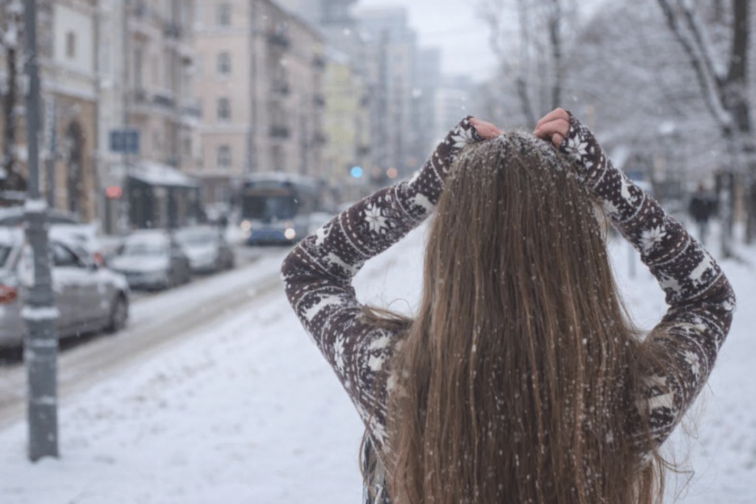 Жінка без шапки йде засніженою міською вулицею взимку