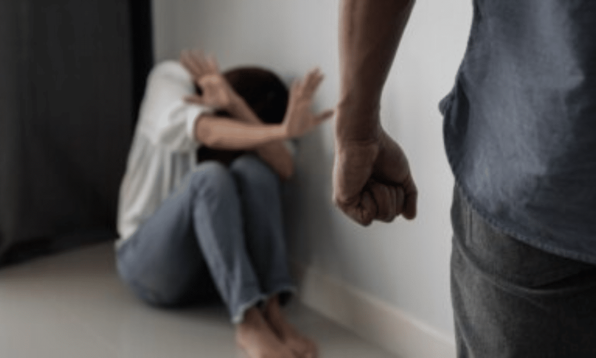 Чоловік з стиснутим кулаком стоїть перед наляканою жінкою, яка сидить у кутку кімнати й закривається руками