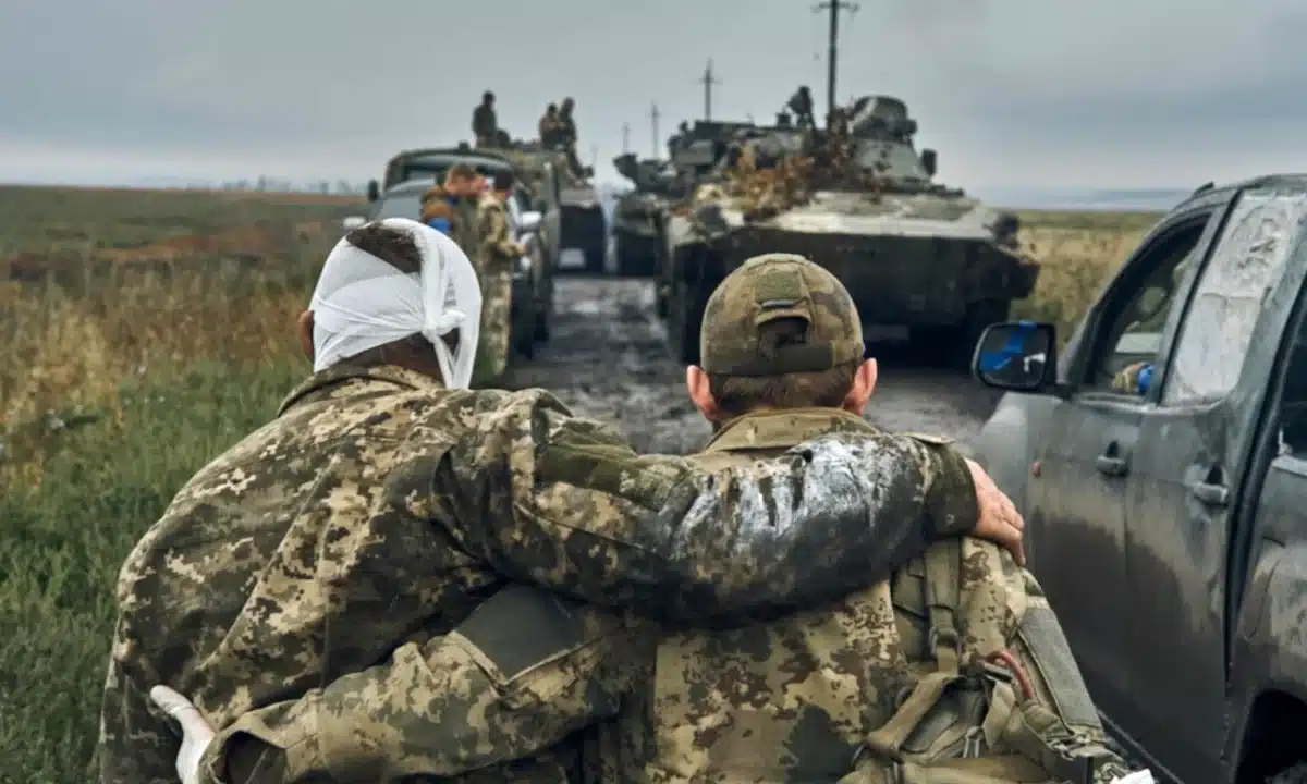 Поранений військовий йде спираючись на плече побратима на фоні військвої техніки.