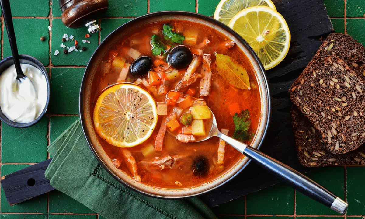 Тарілка солянки з м’ясом, овочами та оливками, подана з часточкою лимона, сметаною та житнім хлібом на дерев’яній дошці.