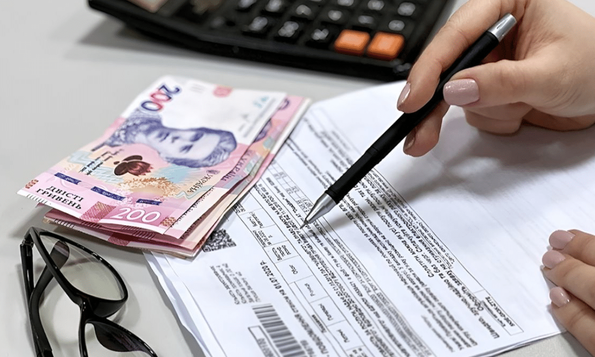 Оформити субсидію у Рівному тепер можна ще за однією адресою Оформлення документів на субсидію: рука з ручкою заповнює бланк, поруч лежать гривневі купюри та калькулятор.