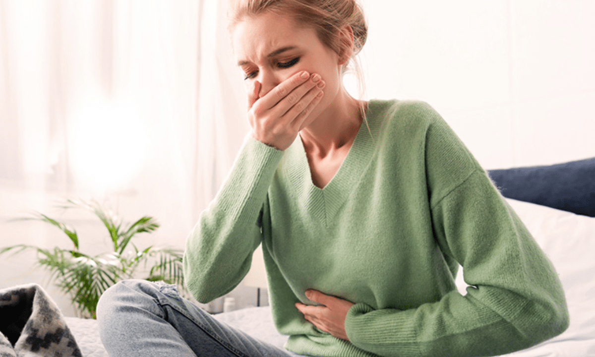 Жінка сидить на ліжку й відчуває нудоту зранку, тримаючись за живіт і прикриваючи рот рукою.