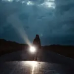 Дівчина йде дорогою вночі з потужним ліхтарем під хмарним небом.